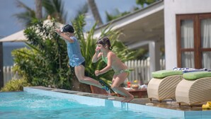 Dos personas saltando a una piscina, con una tumbona y una mesa al fondo.