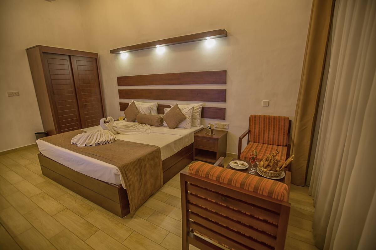 Quarto de hotel com uma cama grande, guarda-roupa de madeira, mesa de cabeceira, uma cadeira e uma pequena mesa com um objeto decorativo.
