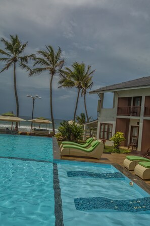 A pool area with lounge chairs, palm trees, and a building with balconies overlooking the ocean. - Avenra Beach Hotel (Hikkaduwa)