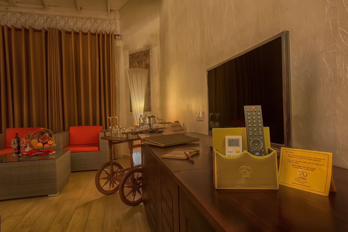Quarto de hotel com piso de madeira, sofá de vime com almofadas vermelhas, uma pequena mesa redonda com uma cesta de frutas, uma cadeira de rodas, uma escrivaninha com um laptop, uma televisão e uma placa na parede.