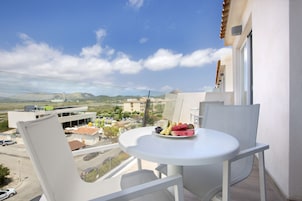 Un balcón con una mesa y sillas blancas, un plato de fruta y vistas a edificios y montañas.