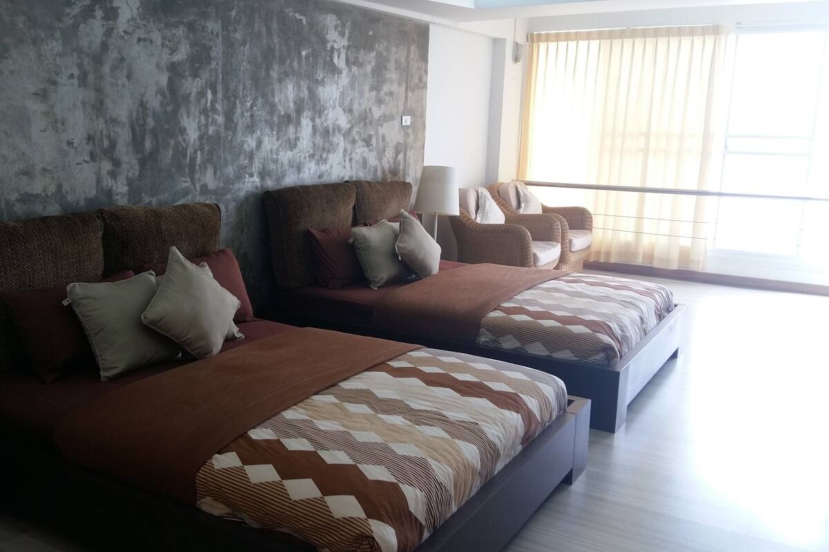 A modern bedroom with two beds, a large window with curtains, and a concrete wall.