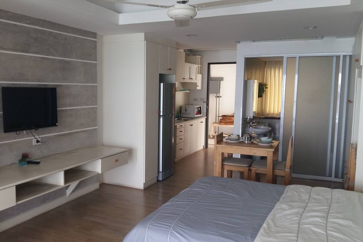 A modern hotel room with a flat-screen TV mounted on a wall, a wooden dining table with chairs, a kitchen area with a refrigerator and microwave, and a bed with a grey and white duvet.