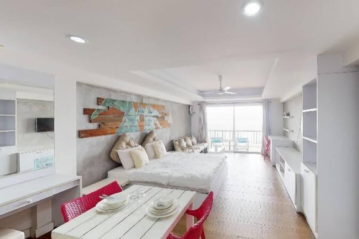 A modern, well-lit room with a large bed, a dining table with chairs, a TV, and a balcony with a view.