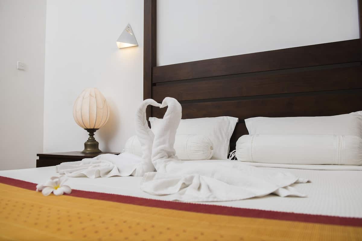 Un lit bien fait, avec du linge de lit blanc et une décoration en forme de cœur réalisée avec des serviettes, une lampe de chevet et un luminaire fixé au mur.