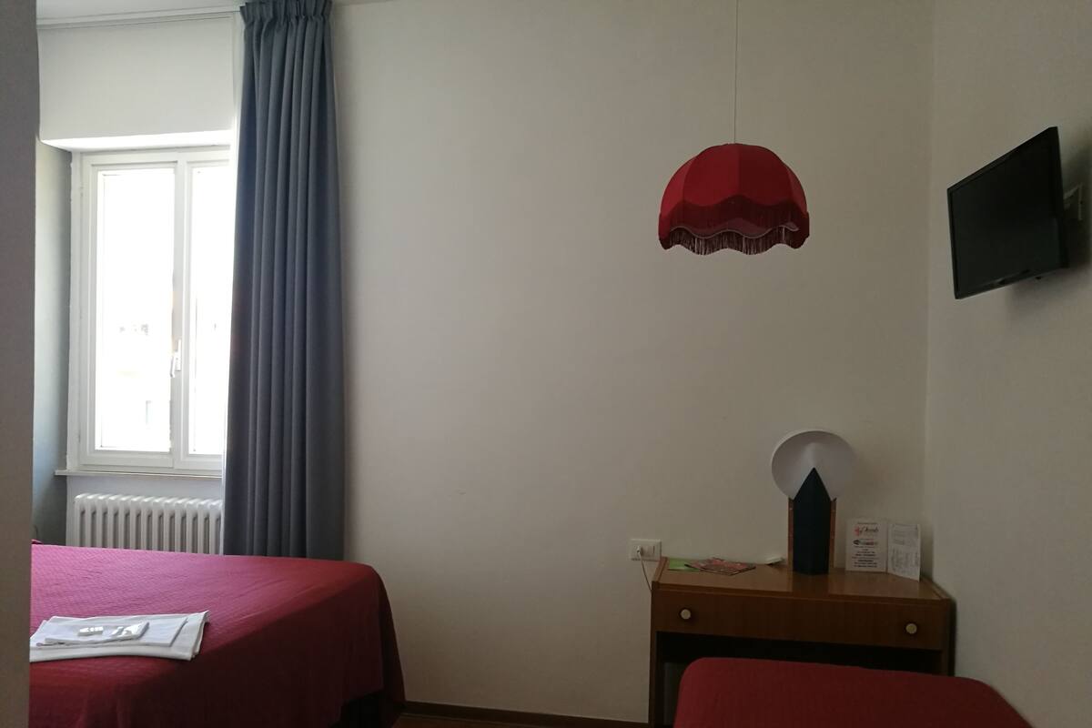 Ein Zimmer mit zwei Betten, einem fenster mit Vorhängen, einem Heizkörper, einem Schreibtisch mit Lampe und einem an der Wand befestigten Fernseher.