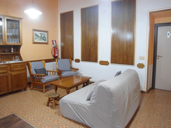 Una sala de estar con un sofá, dos sillones, una mesita de centro de madera, un taburete pequeño, un armario y un extintor en la pared.