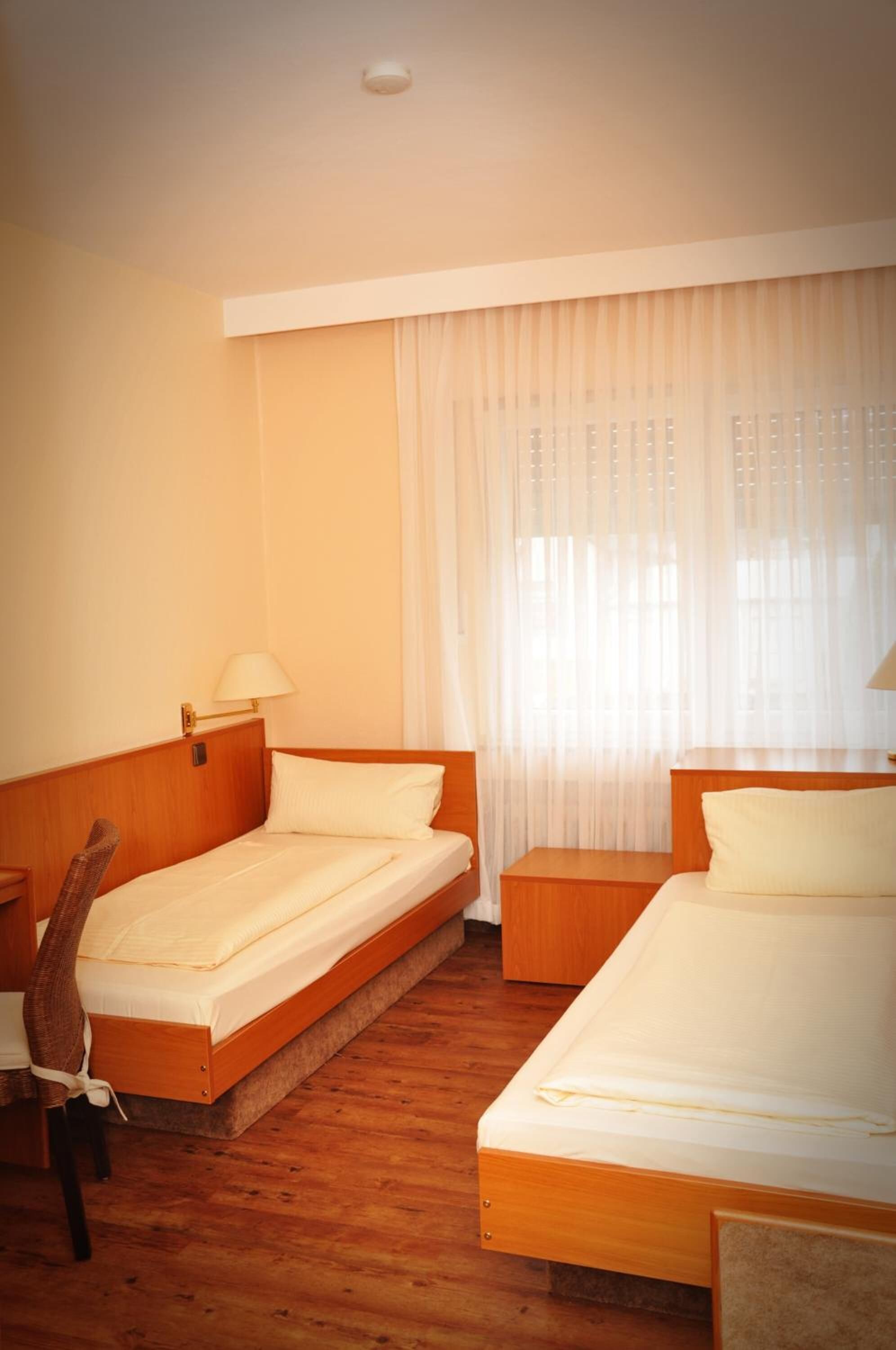 A hotel room with two single beds, a wooden headboard, a wooden nightstand, a chair, and a window with sheer curtains.