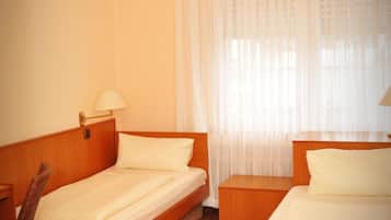 Habitación de hotel con dos camas individuales, cabecera y mesita de noche de madera, una silla y una ventana con cortinas traslúcidas.