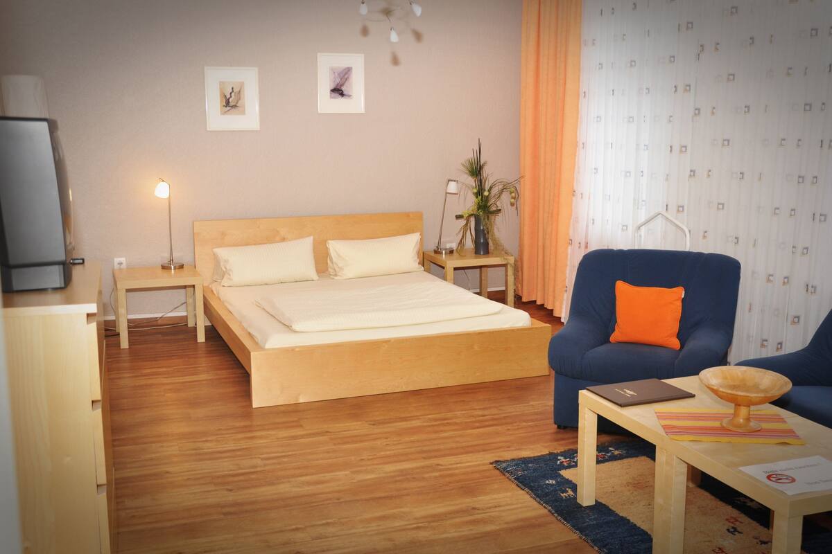 A hotel room with a wooden bed, a blue armchair, a small table with a laptop, and a bedside lamp.