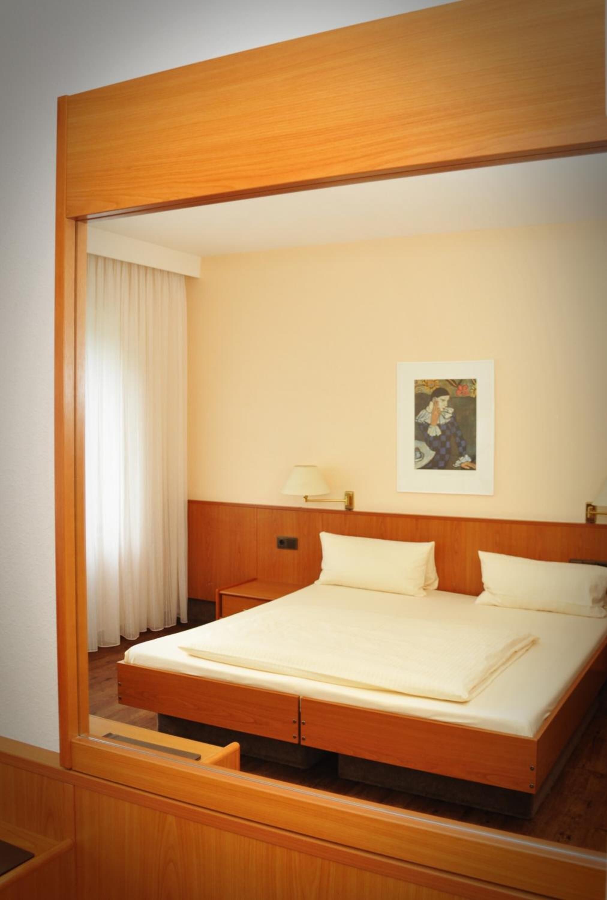 A hotel room with a wooden bed frame, a single bedside table with a lamp, a framed picture on the wall, and a window with curtains.