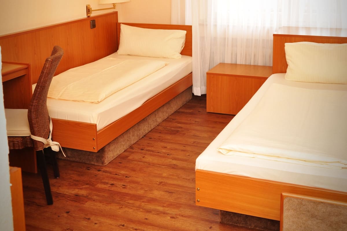 A hotel room with two single beds, a wooden headboard, a desk with a chair, and a window with sheer curtains.