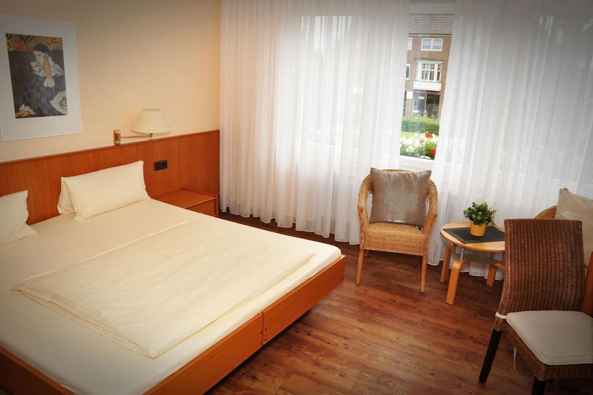 A hotel room with a large bed, two chairs, a small table with a plant, and a window with sheer curtains.