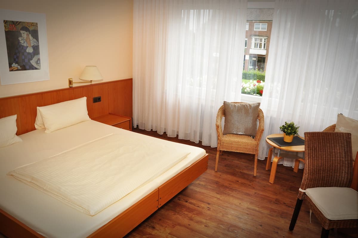 A hotel room with a large bed, a chair, a small table with a plant, and a window with sheer curtains.