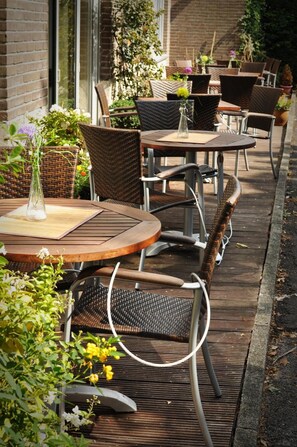 An outdoor patio with round tables and wicker chairs, surrounded by potted plants and flowers.