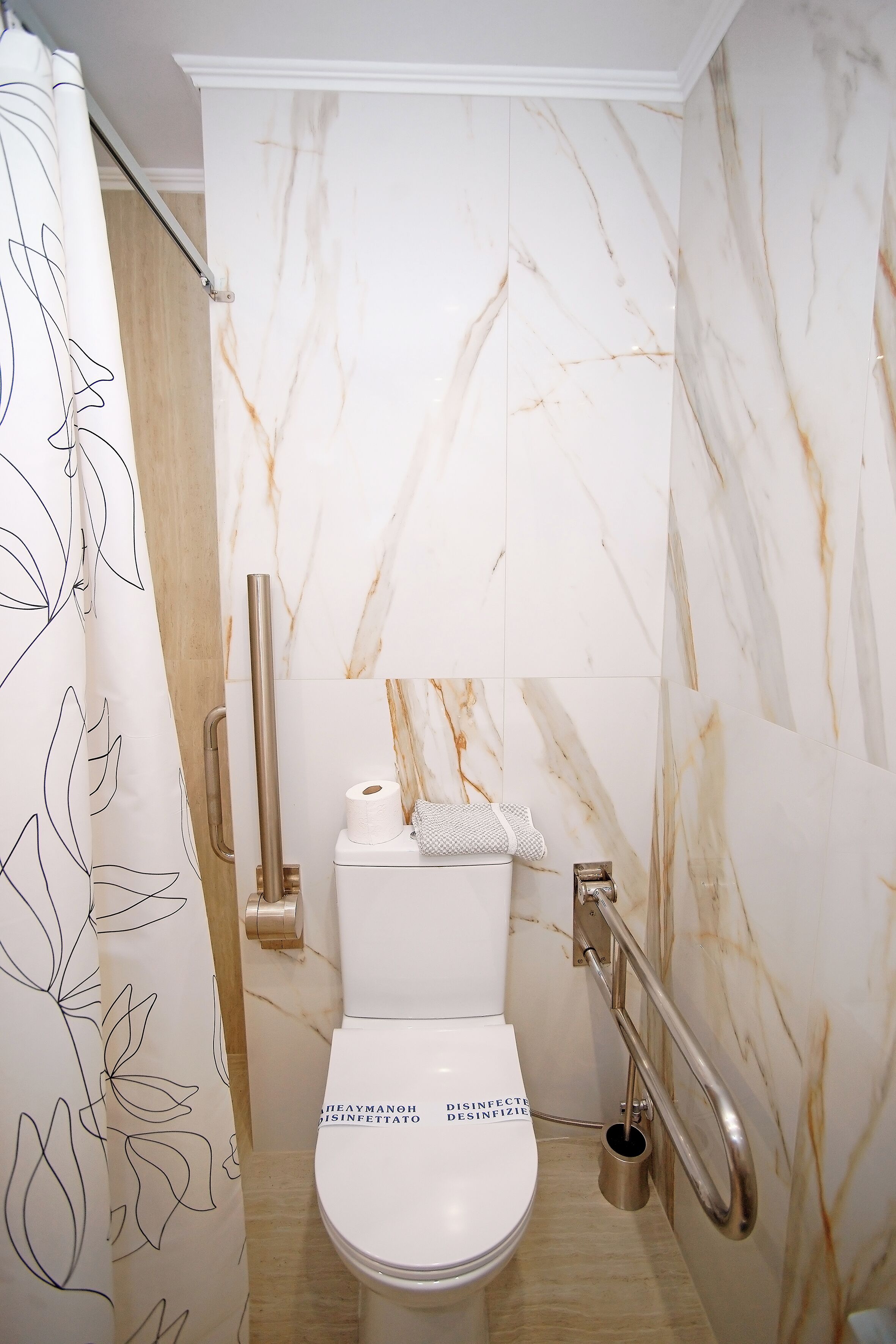 Baño con inodoro, barras de apoyo, un rollo de papel higiénico y un pequeño cubo de basura.