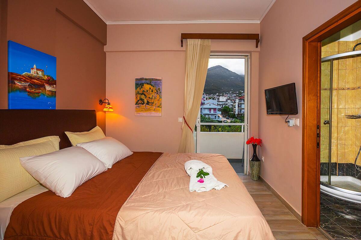 A hotel room with a bed, a painting, a TV, and a view of a town through a window.