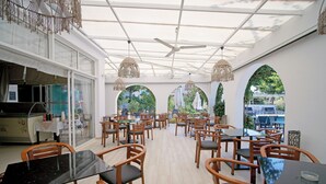 Un restaurante con varias mesas y sillas, un ventilador de techo y grandes ventanales que dan a zonas verdes.