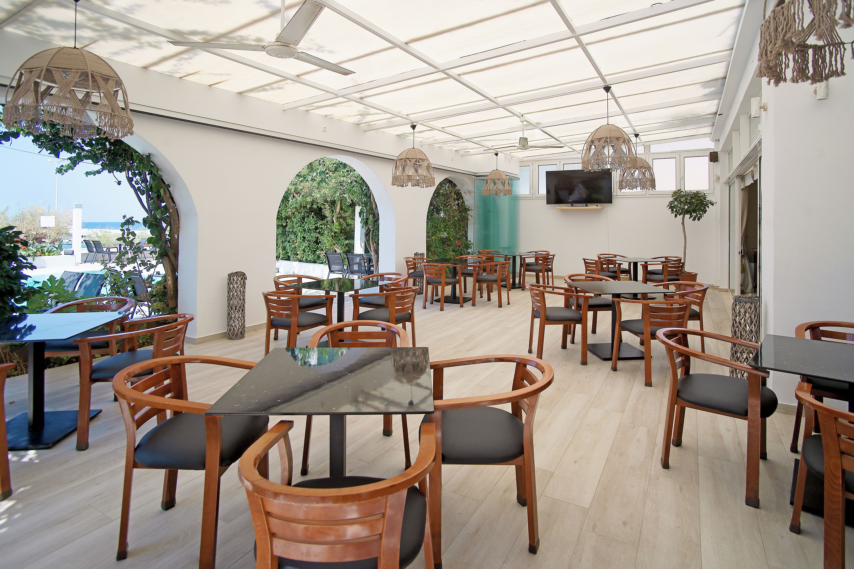 A spacious indoor dining area with multiple wooden tables and chairs, a large TV screen on the wall, and a view of the outdoors through large windows.