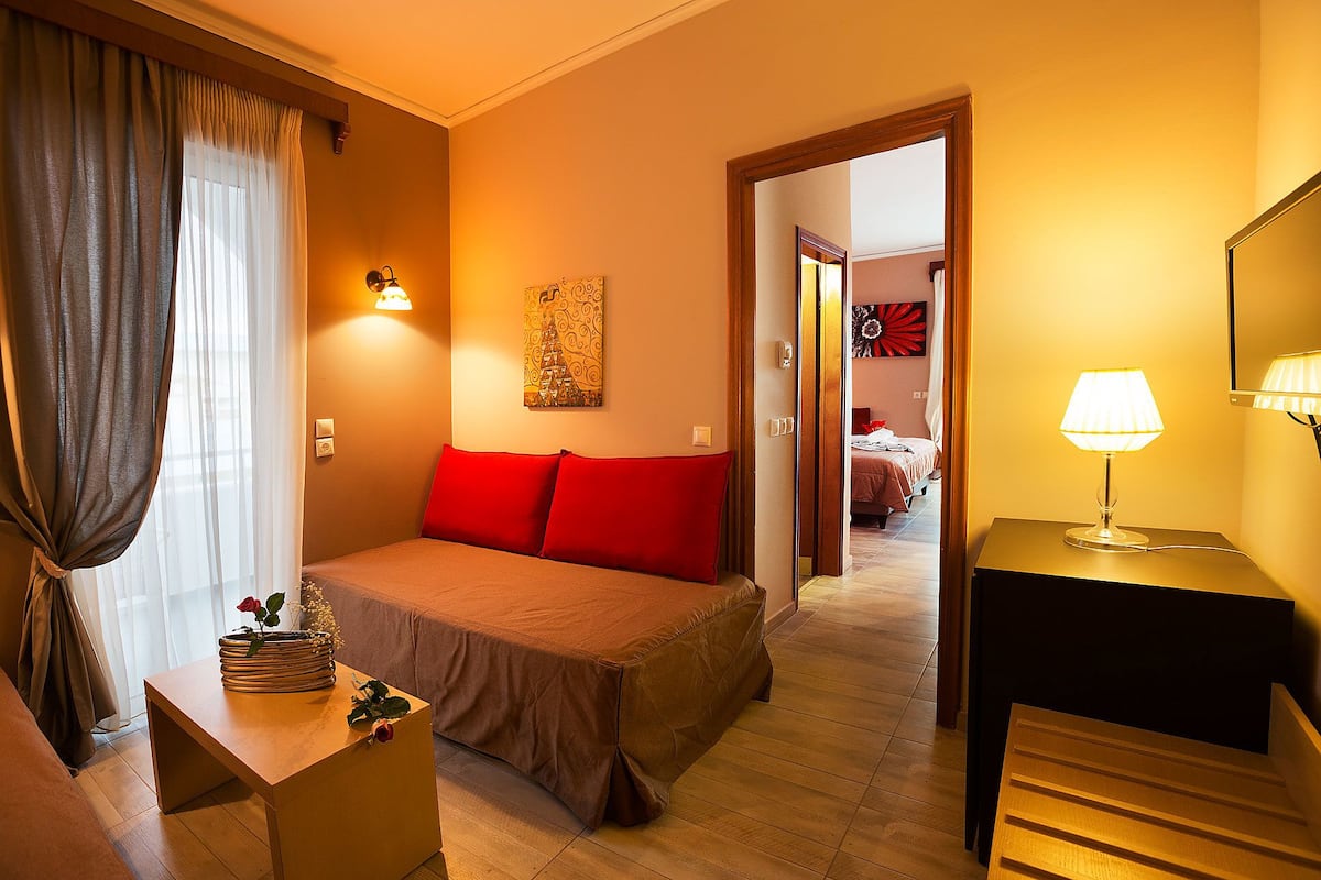 A hotel room with a bed, a bedside table with a lamp, a painting on the wall, and a view into the adjacent room.