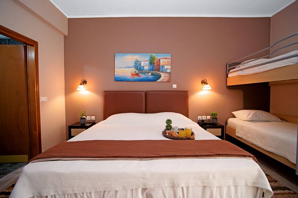 A hotel room with a large bed, a smaller bunk bed, two bedside tables with lamps, a painting on the wall, and a tray with breakfast items.