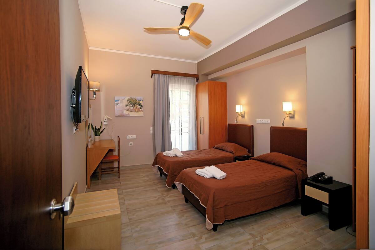 A hotel room with two beds, a desk, a chair, a ceiling fan, and a TV.