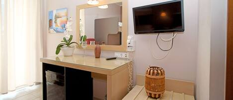 A compact room with a wooden desk, a small refrigerator, a television mounted on the wall, and a mirror above the desk.