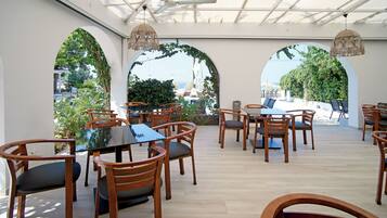 A covered outdoor dining area with wooden tables and chairs, large arched windows, and hanging rattan lamps.