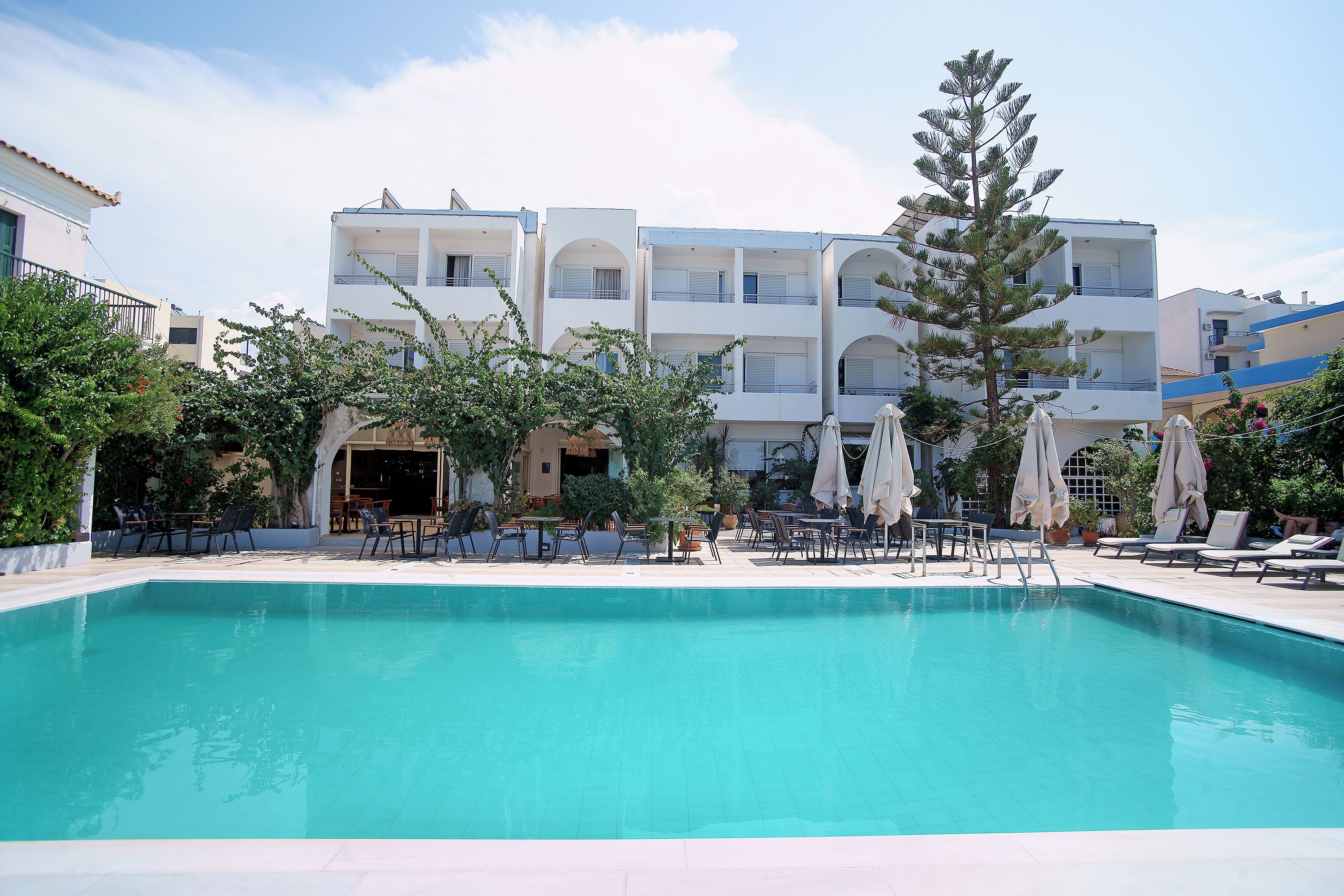 A swimming pool with lounge chairs and umbrellas, surrounded by buildings with balconies and a clear sky.