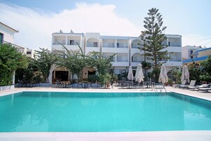 Una piscina con sillones de descanso y sombrillas, rodeada de edificios con balcones y un cielo despejado.