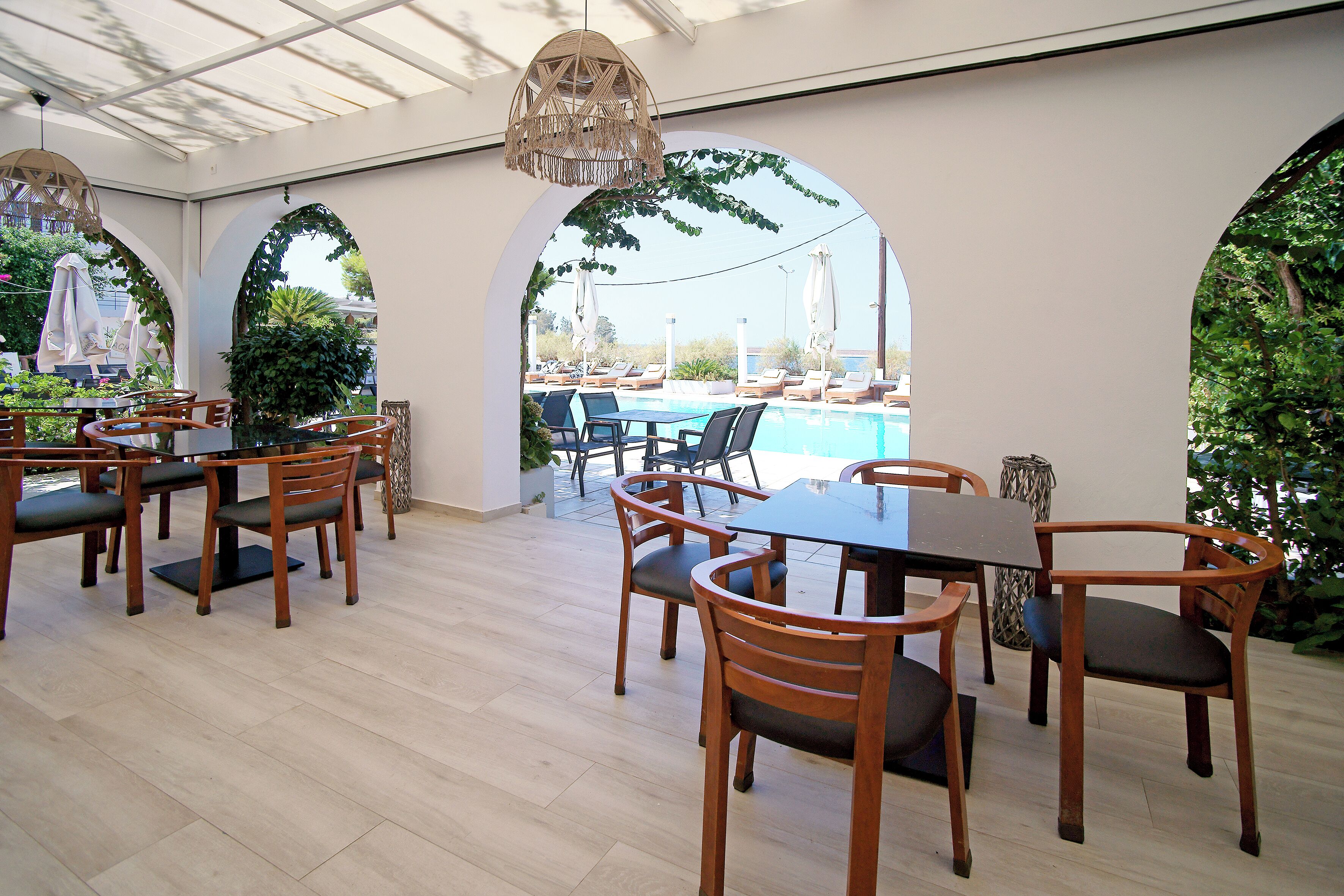 Un'area pranzo all'aperto coperta, con tavoli e sedie in legno, una piscina sullo sfondo e porte ad arco.
