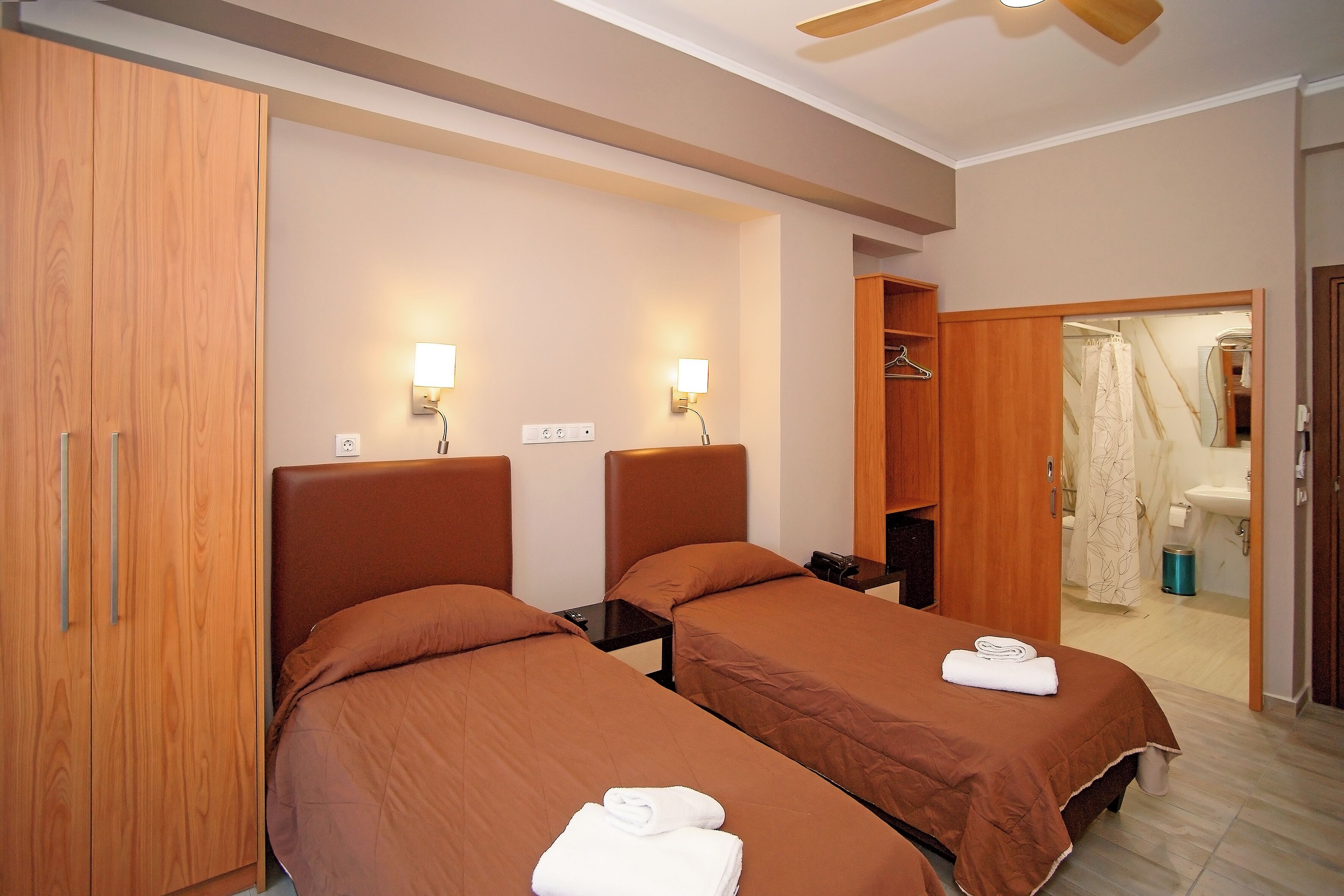 Habitación de hotel con dos camas, un armario de madera, lámparas de noche y un baño visible a través de una puerta abierta.