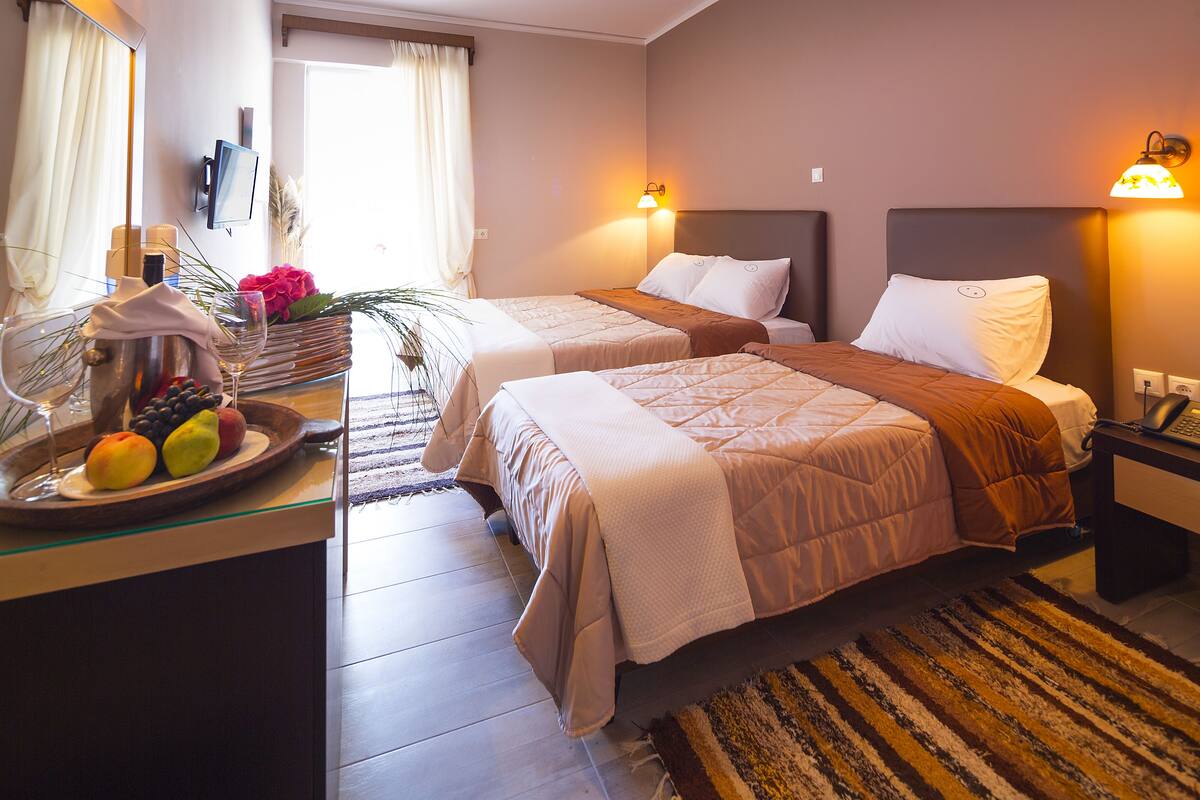 A hotel room with two beds, a bedside table with a fruit bowl, a lamp, and a view of the outdoors.
