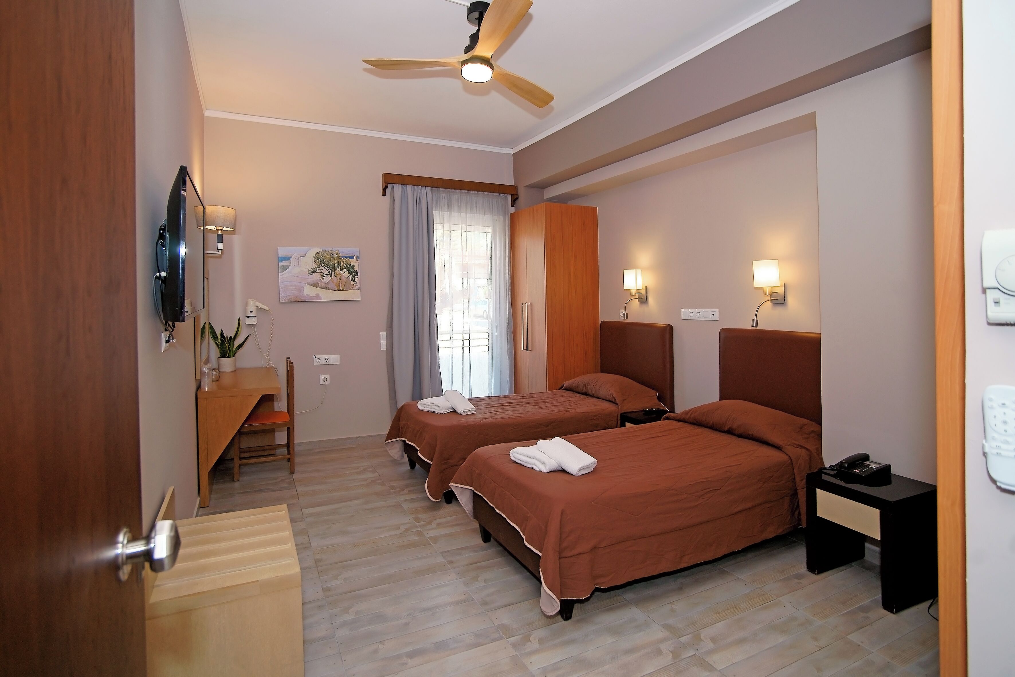 Habitación de hotel con dos camas, un escritorio, una silla, un ventilador de techo y una ventana con cortinas.
