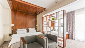 A hotel room with a large bed, two armchairs, a wooden headboard, a built-in bookshelf, and a patterned carpet. - AdvaStay by KING's (Munich)