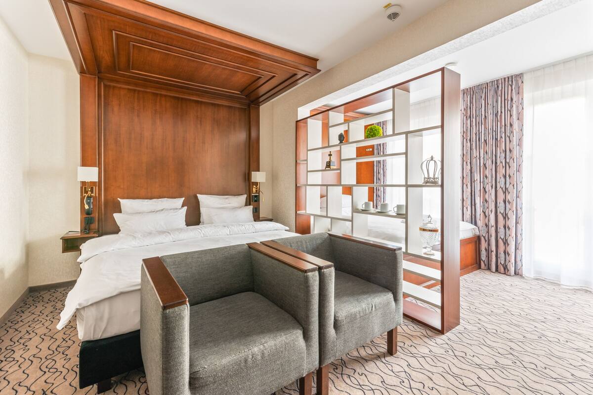 A hotel room with a large bed, two armchairs, a wooden headboard, a built-in bookshelf, and a patterned carpet.