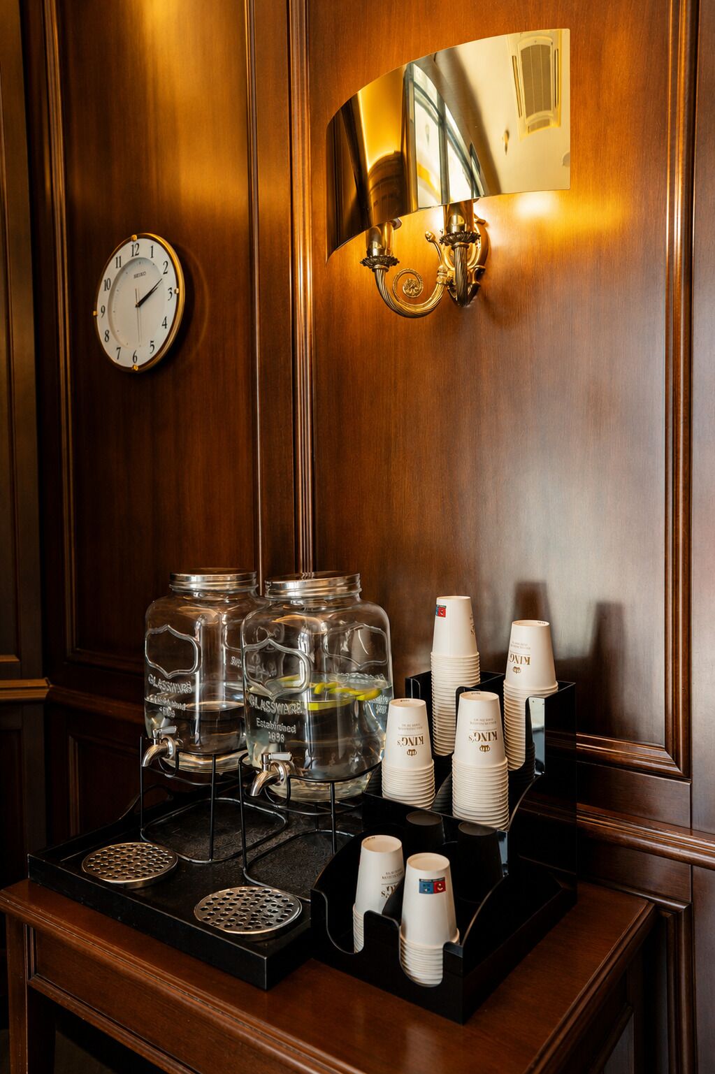 Ein Hotel-Empfangsbereich mit einer Wand aus Holzpaneelen, einer Wanduhr, einem Spiegel und einer Kaffeestation mit Tassen und Wasserspender.