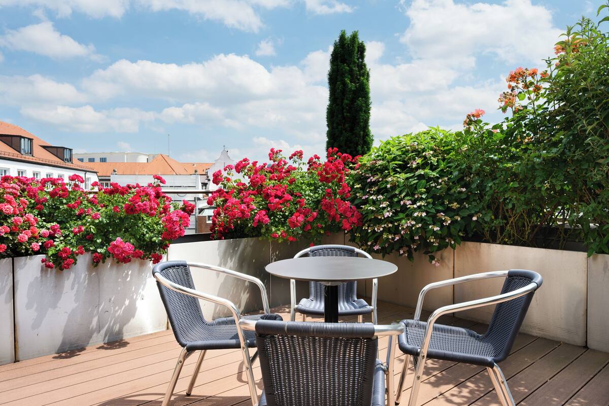A rooftop terrace with a round table and four chairs, surrounded by lush greenery and vibrant flowers.
