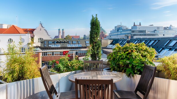 A rooftop terrace with a round table and two chairs, surrounded by potted plants and overlooking a cityscape with modern buildings and a church tower. - AdvaStay by KING's (Munich)