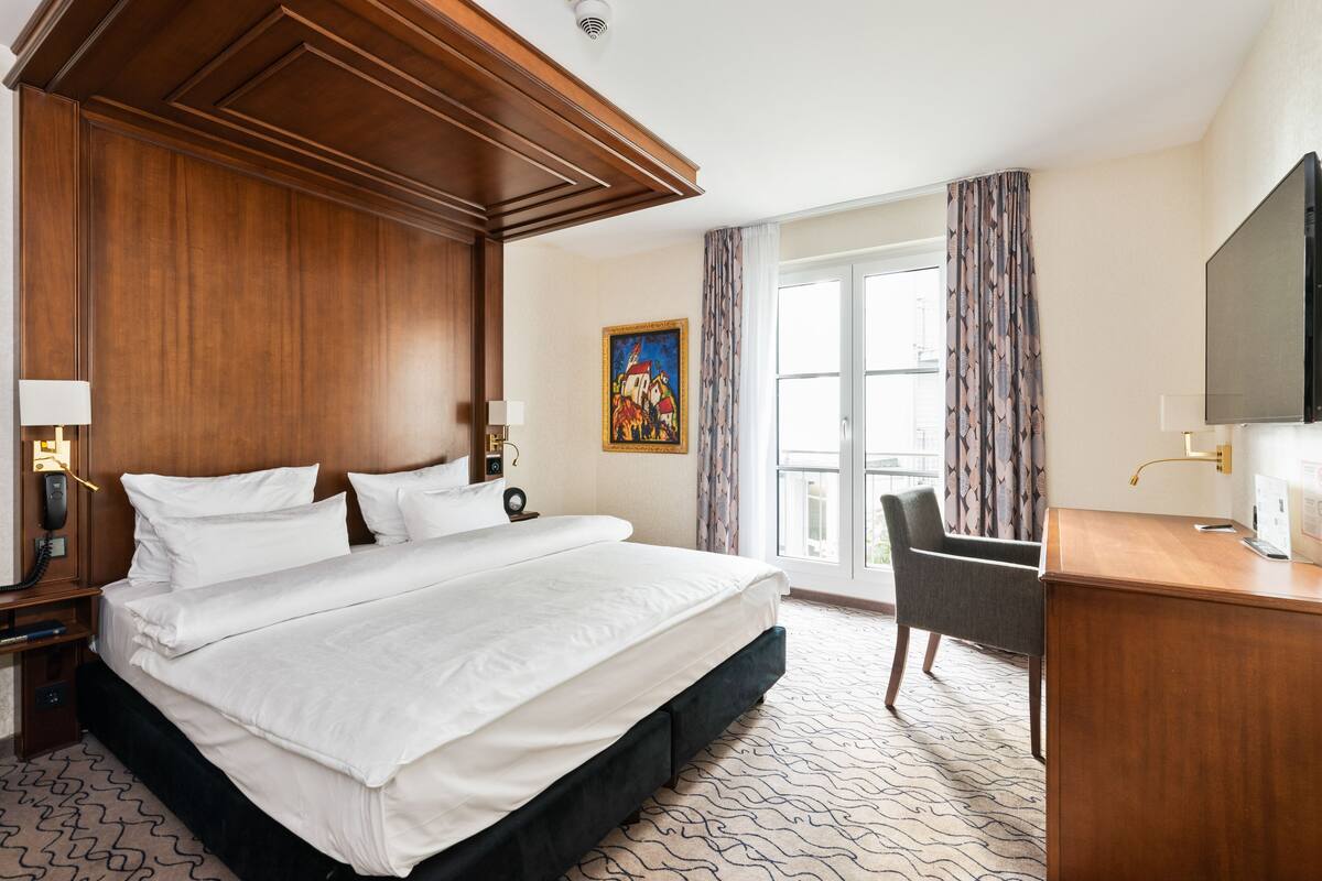 A hotel room with a large bed, a desk, a chair, a television, and a painting on the wall.