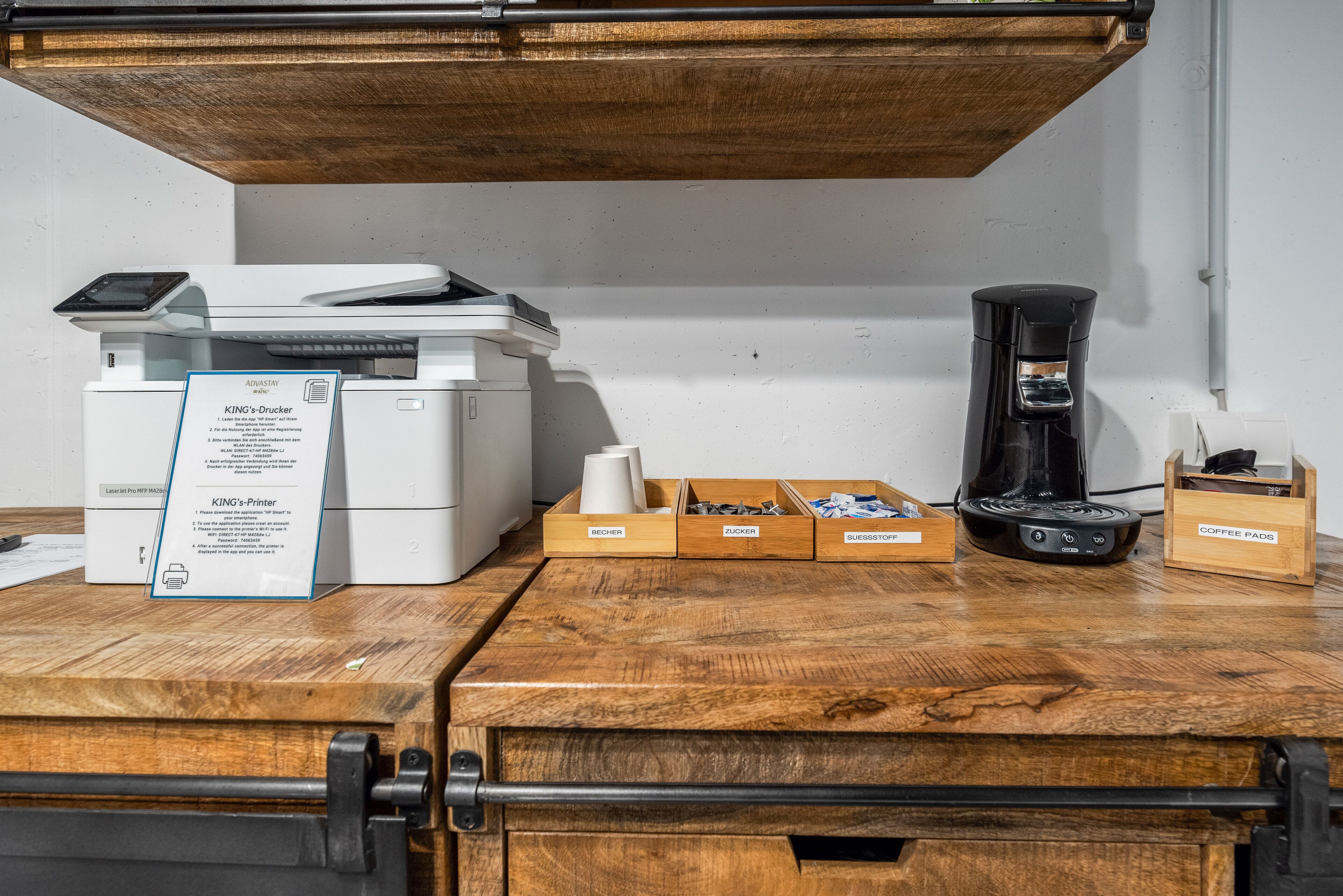 A printer, coffee maker, and boxes labeled "ROCHER," "ZUCKER," and "SUSSTOPP" on a wooden counter.