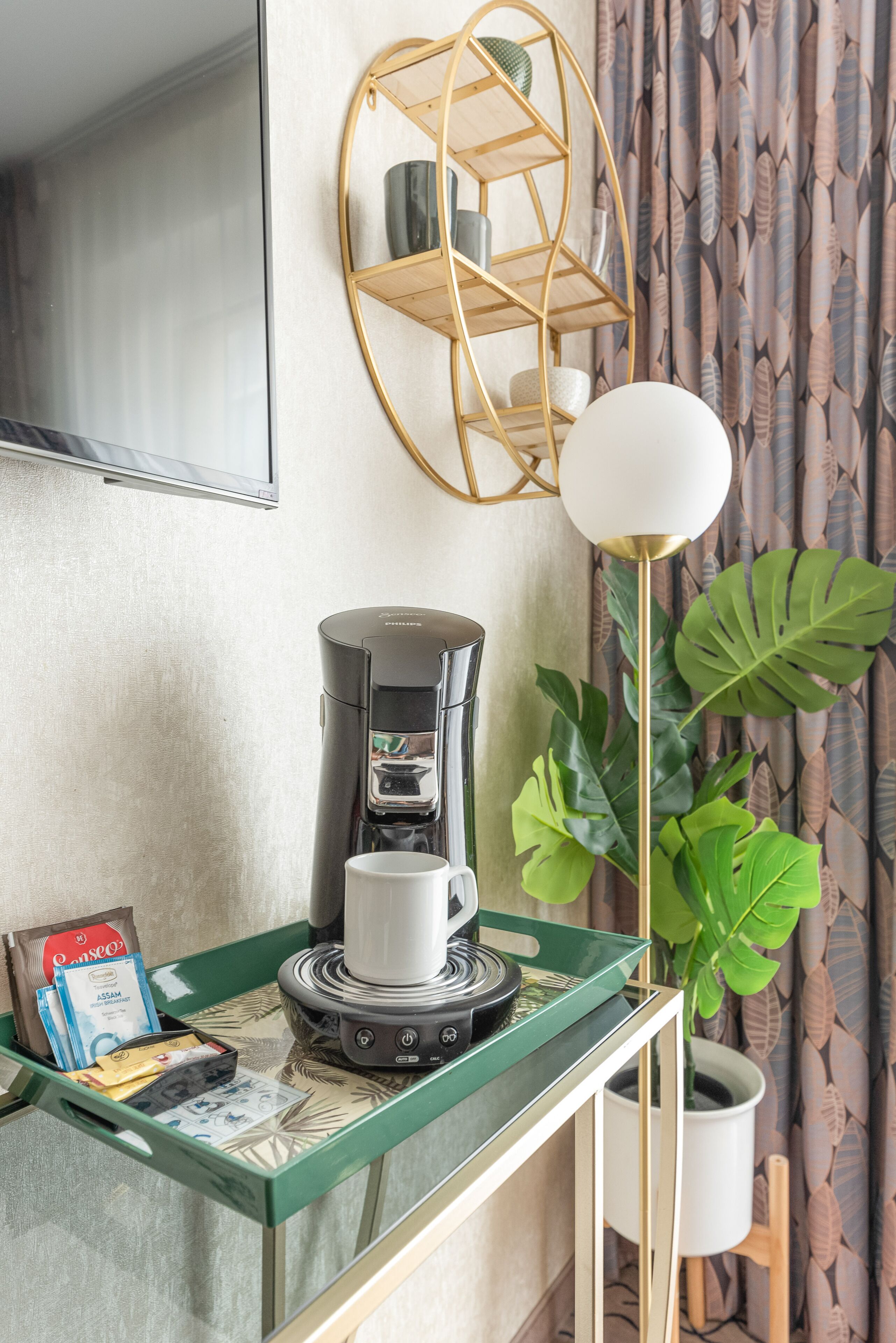 A coffee machine on a table with a cup, a tray of coffee pods, and a plant.