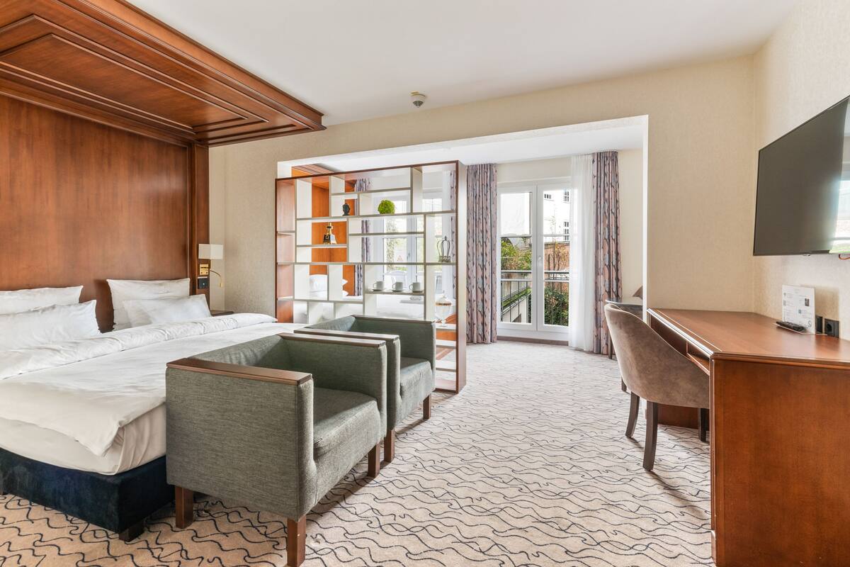 A hotel room with a bed, two armchairs, a desk, and a television. There is a large window with a view of the outdoors.