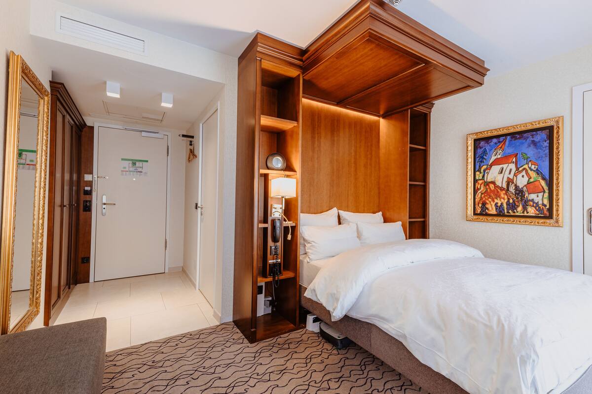 A hotel room with a bed, a wardrobe, a desk, and a painting on the wall.