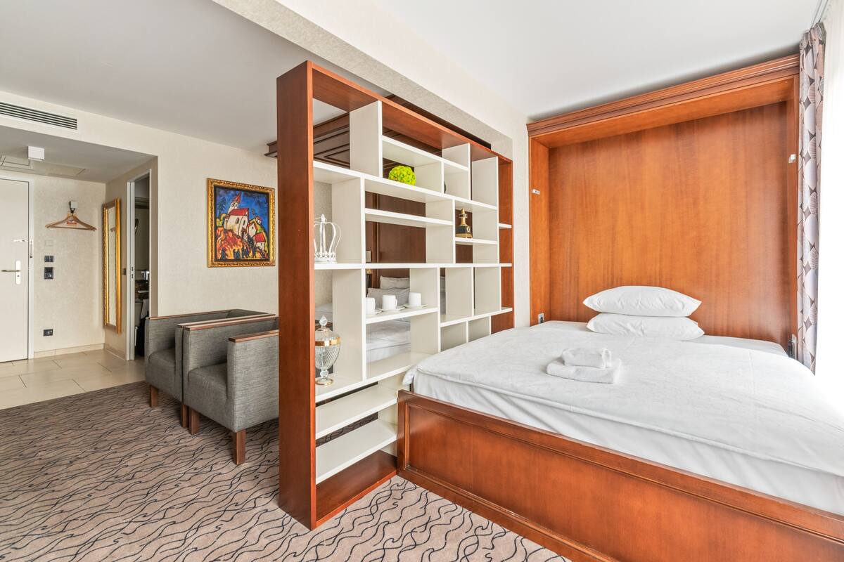 A hotel room with a large bed, a wardrobe with multiple shelves, a seating area with two chairs, and a painting on the wall.