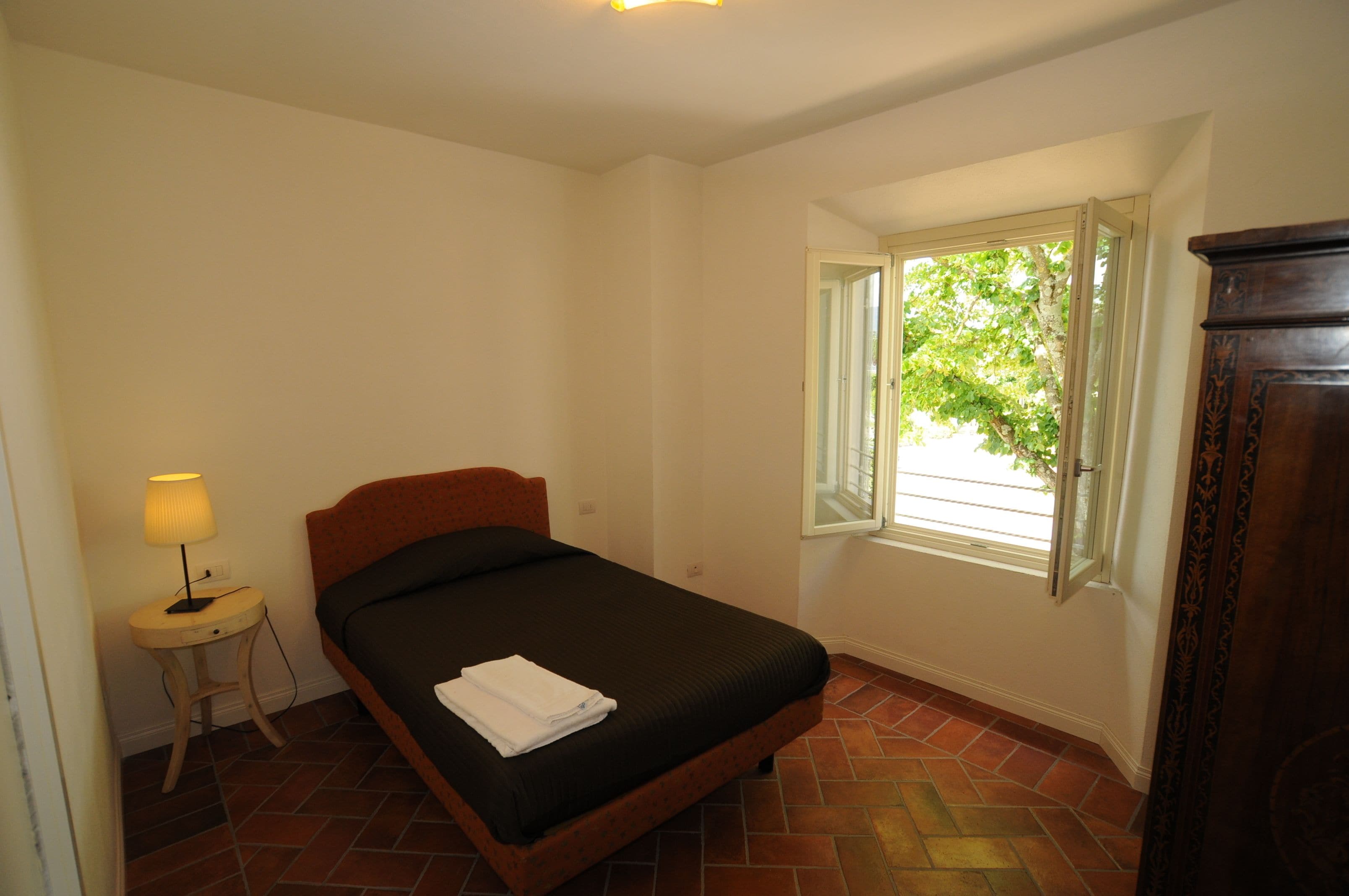 Une chambre avec un lit simple, une table de chevet avec une lampe, une fenêtre donnant sur un paysage verdoyant et une armoire en bois.