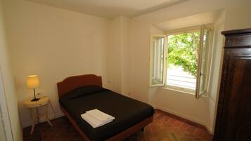 Une chambre avec un lit simple, une table de chevet avec une lampe, une fenêtre donnant sur un paysage verdoyant et une armoire en bois.