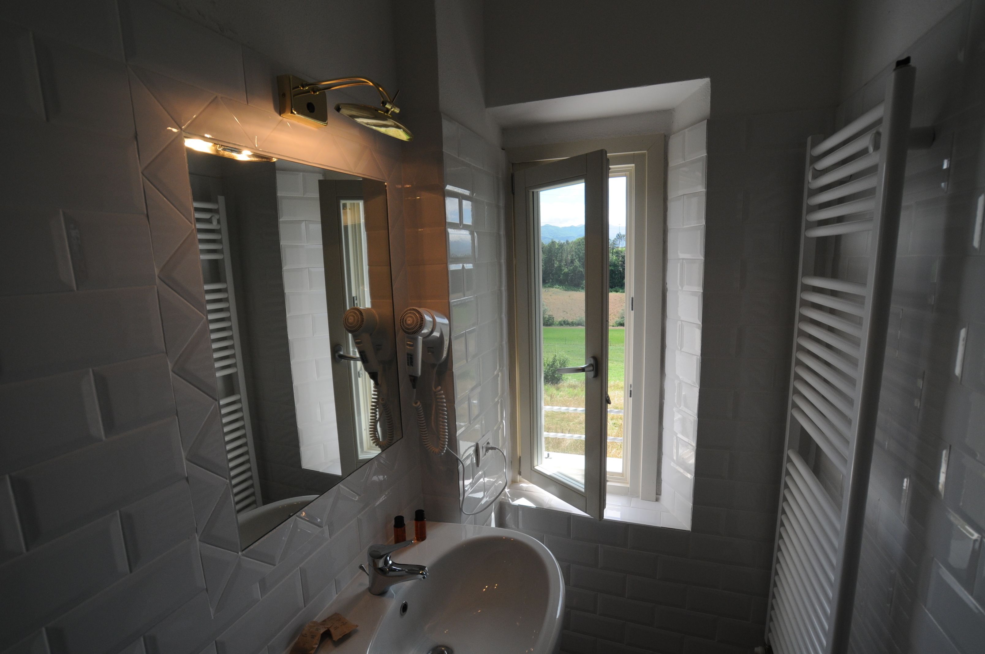 Casa de banho com lavatório branco, espelho e janela com vista para vegetação.