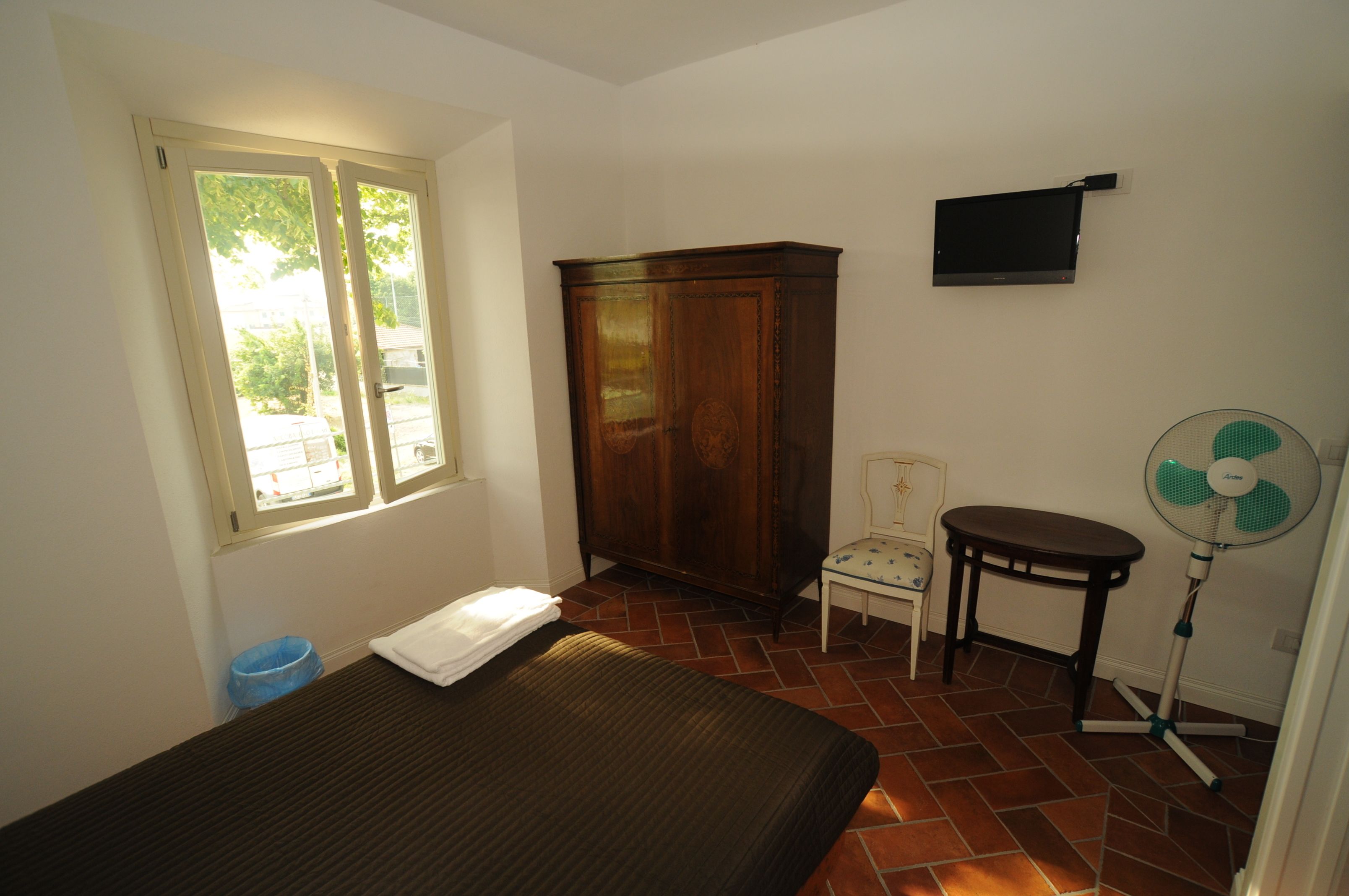 A room with a bed, a wooden wardrobe, a chair, a small table, a standing fan, and a window with a view of the outside.
