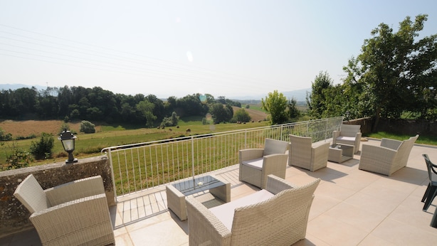 A patio with wicker furniture and a glass table, overlooking a green landscape with trees and hills.
